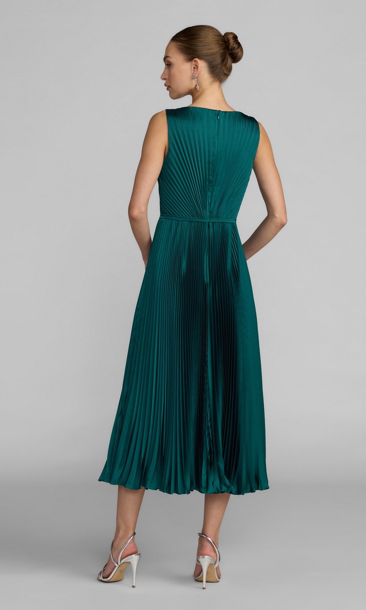 A woman stands with her back to the camera, wearing the Mac Duggal 56326 midi party dress—a sleeveless, emerald green pleated charmeuse piece with an A-line skirt—paired with white high-heeled sandals.
